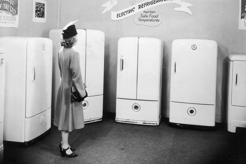 A vintage black-and-white photograph shows a woman looking at display model refrigerator.