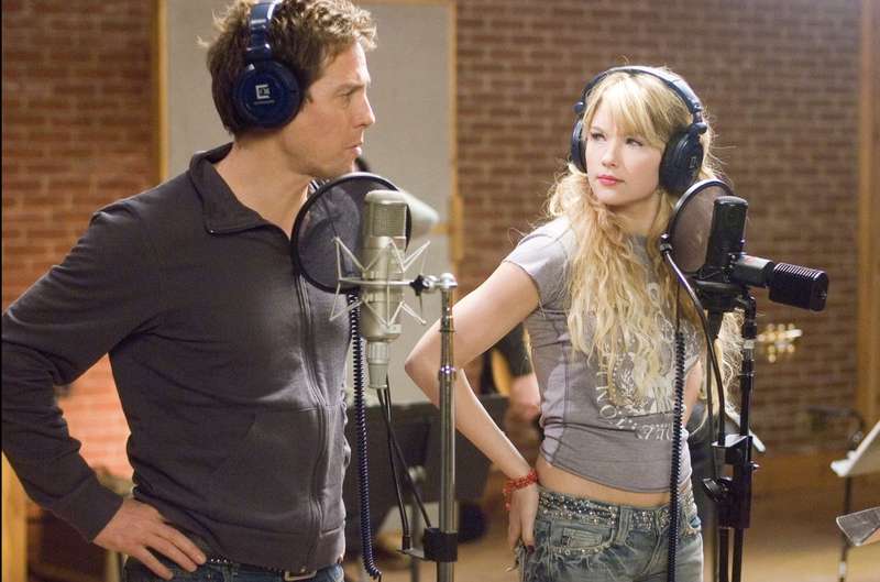A man and a woman in a sound studio standing at microphones and looking at each other.