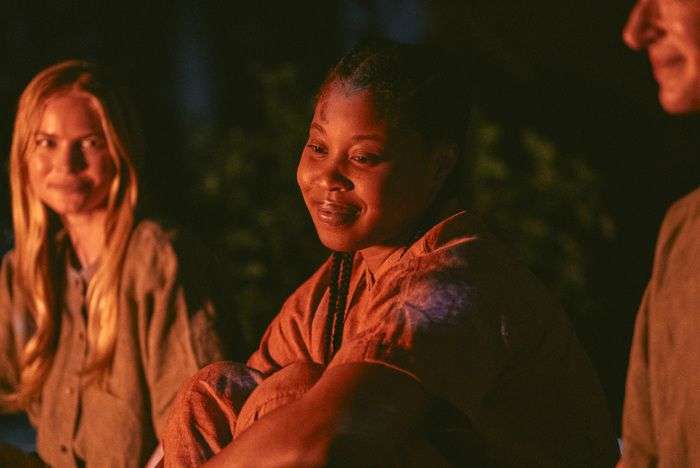 A young black woman smiling in the glow of a campfire as another young woman looks at her.