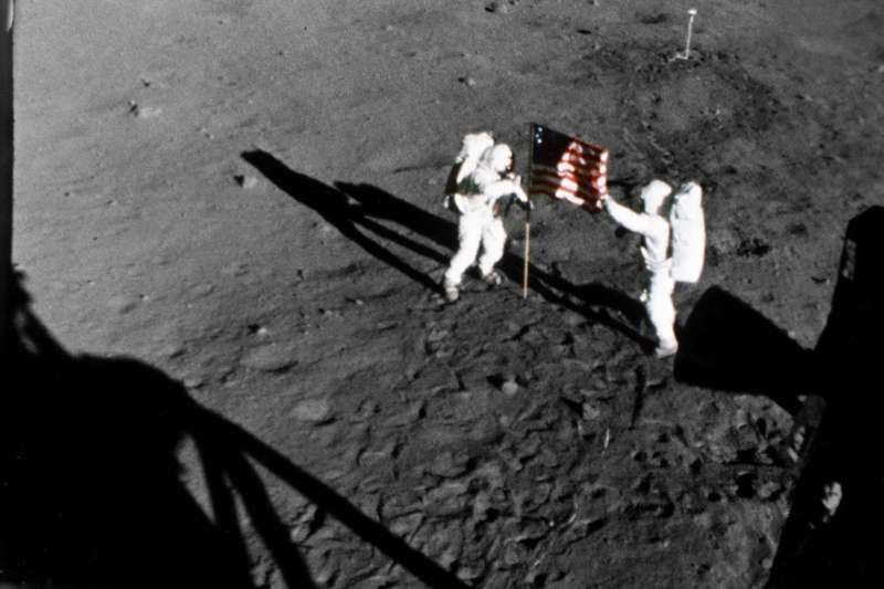 Two astronauts on the lunar surface stand on either side of the American flag on a pole planted in the dirt.