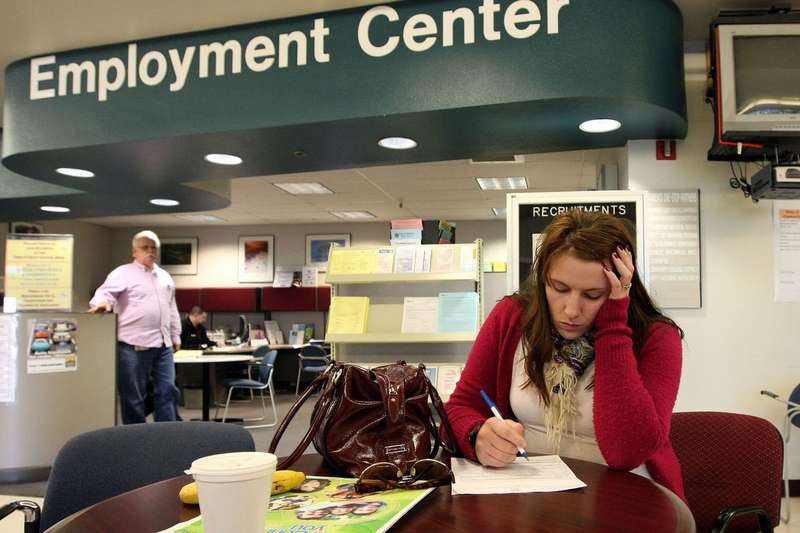 A woman at a table under a sign that reads “Employment Center” sits and fills out a form with her head in one hand.